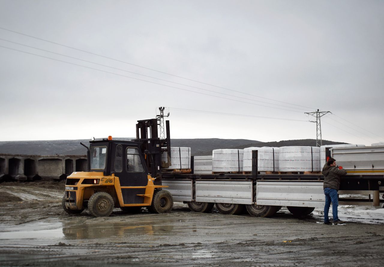 gallery-04 A forklift is loading cargo onto a truck in a muddy logistics area with a worker assisting.