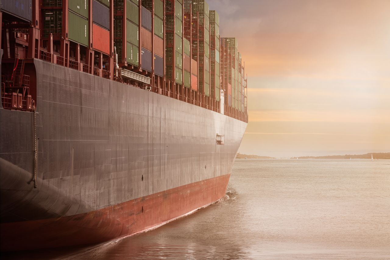 services-01 A colossal cargo ship loaded with containers navigates through calm waters against a vibrant sunset sky.