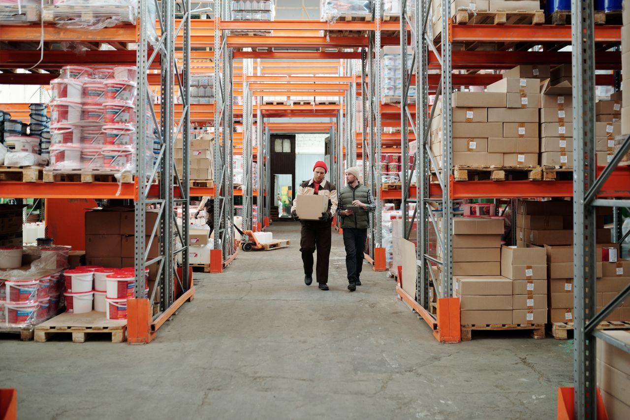 services-02 Two workers handle a package in a spacious warehouse surrounded by shelves stocked with boxes and products.